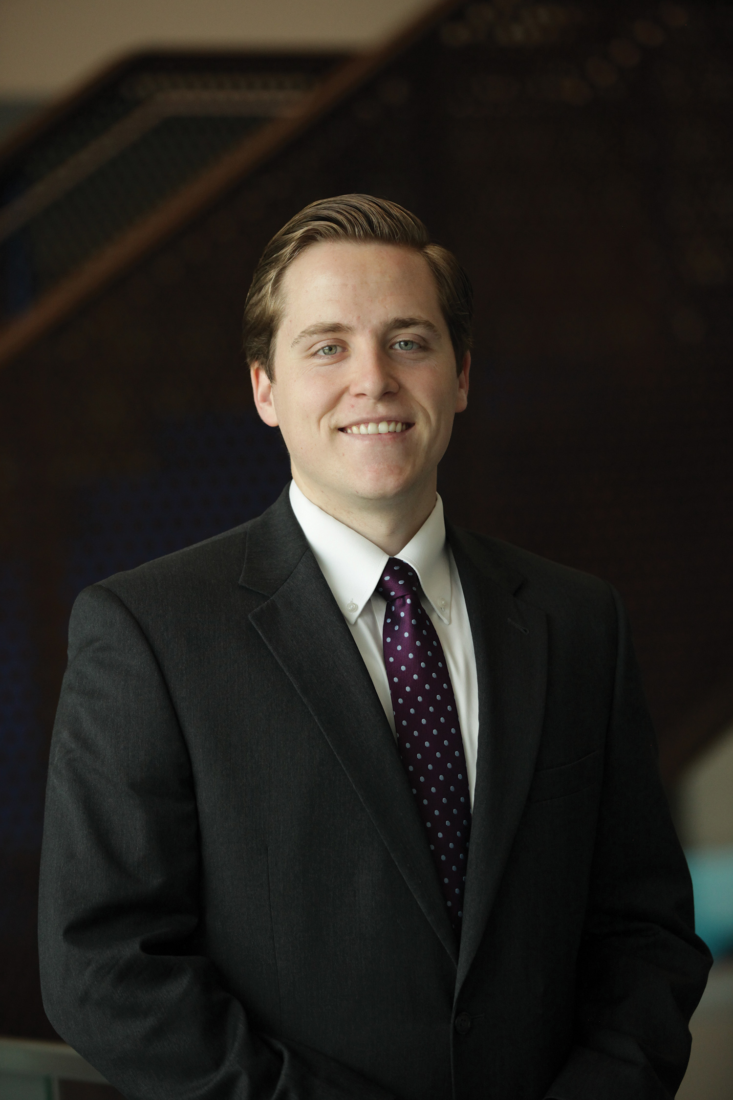 Malcolm Proudfit wears a white shirt, a purple tie with white polka dots, and black jacket while smiling near the stairs in Capitol Federal Hall.