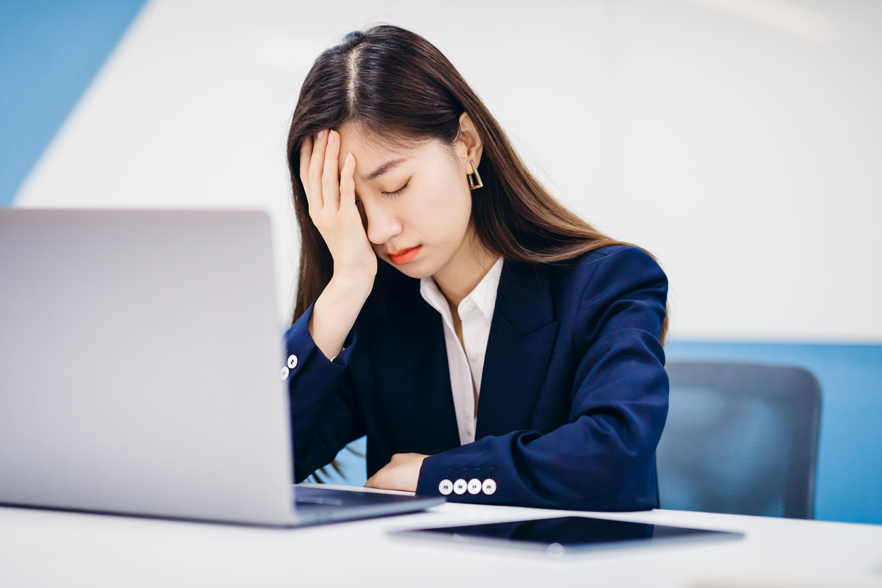 A woman seated in front of a laptop holds her head with her eyes closed