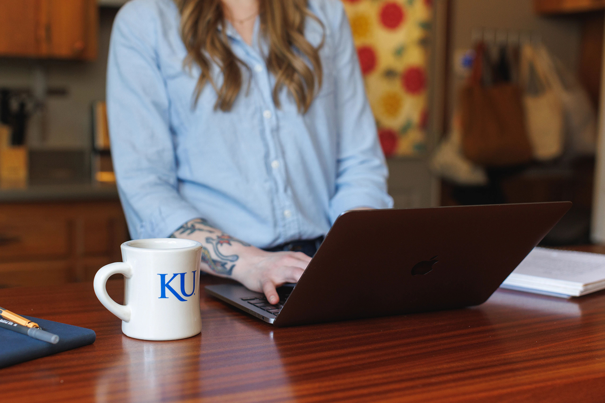 A woman at a counter uses a laptop with a KU mug next to the laptop